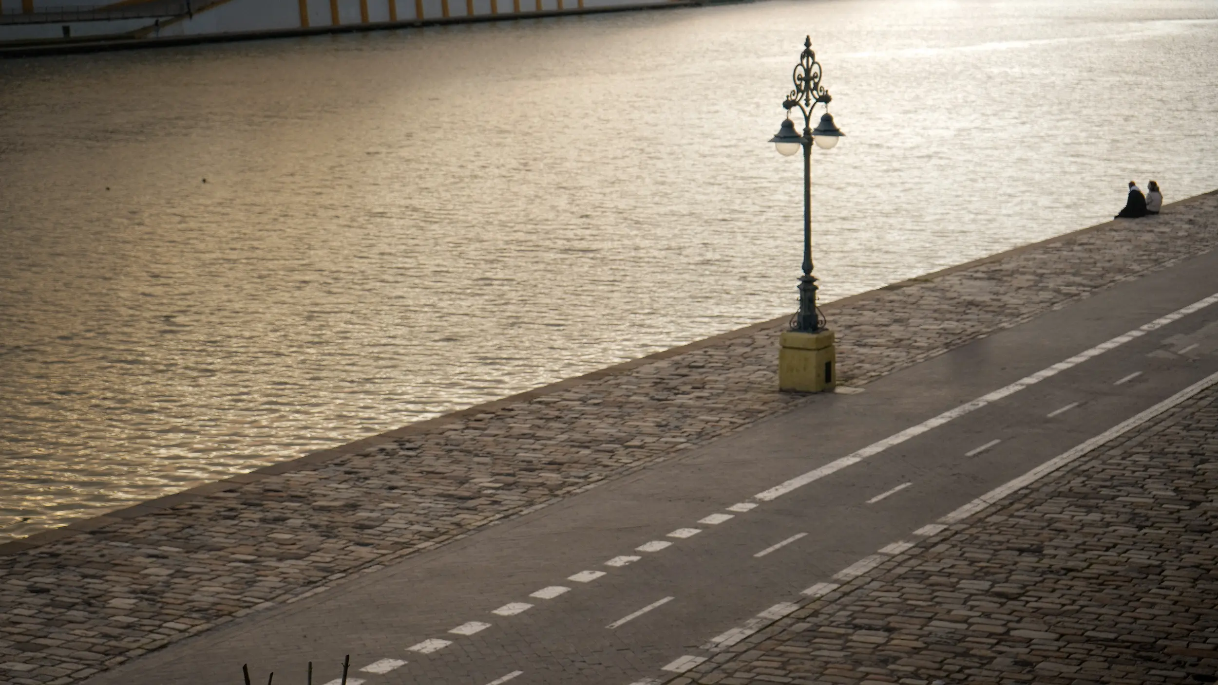 Fotografía urbana del paseo junto al río Guadalquivir en Sevilla con carril bici, farolas y una pareja descansando
