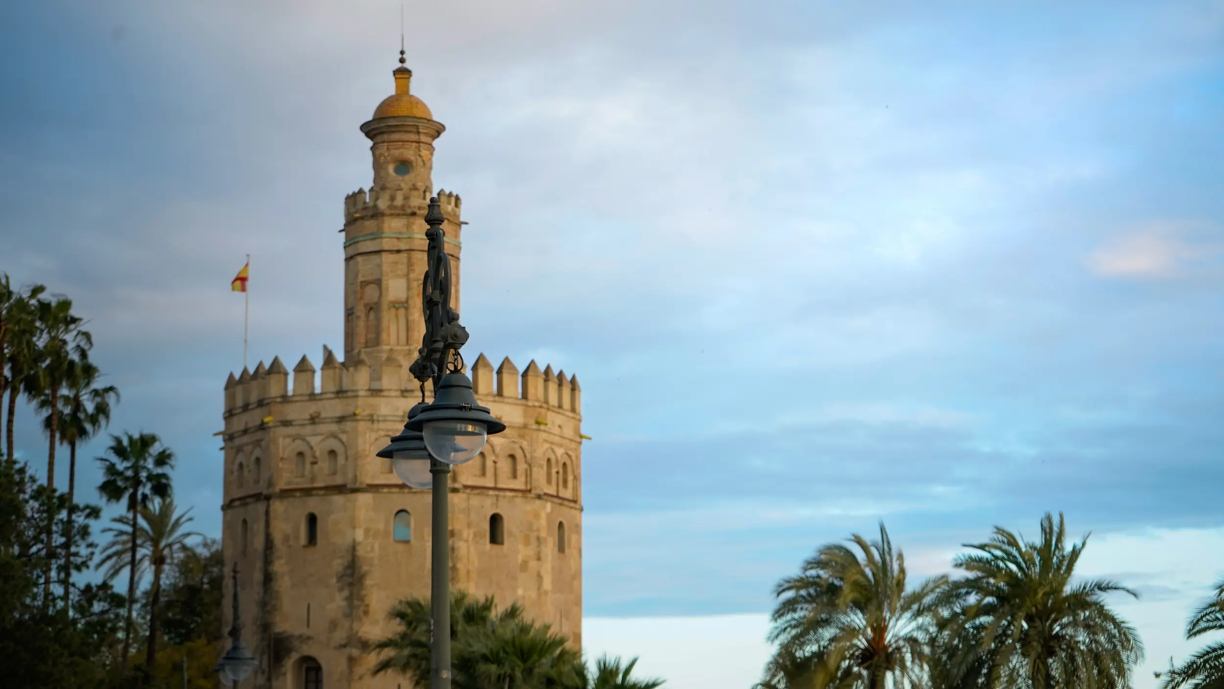 Paisaje urbano de la icónica Torre del Oro en Sevilla enmarcada por palmeras y una farola clásica delante